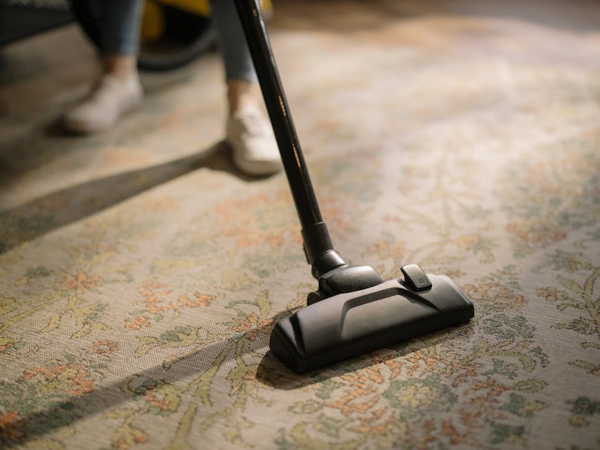 A person kneeling on a patterned area rug in a residential room, working with a yellow and black carpet cleaning machine. They are opening the machine’s lid, revealing the internal components, while