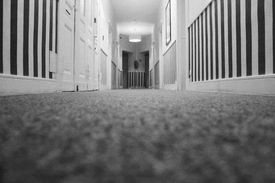 A person using a vacuum cleaner with a black extension hose on an ornate, patterned carpet in a room with wooden flooring. The individual is wearing casual clothing, including beige shoes and dark jea