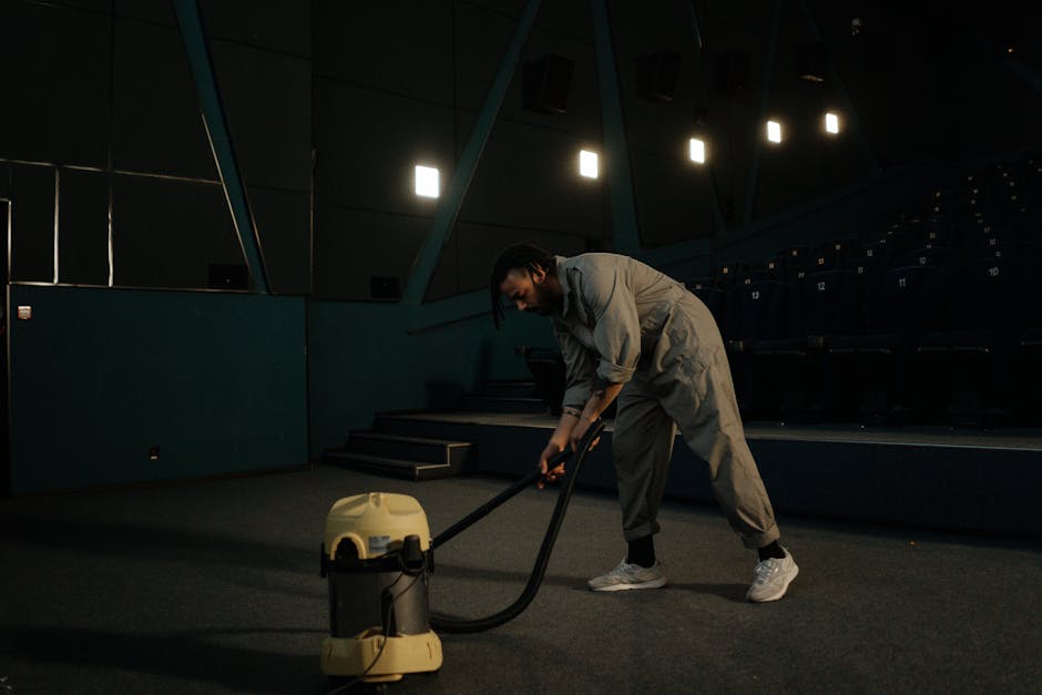 A professional cleaner from Carpet Cleaning Bromley, dressed in full white protective suit, gloves, and face mask, is using a steam cleaning machine to deep clean a beige carpet in a bright, modern li