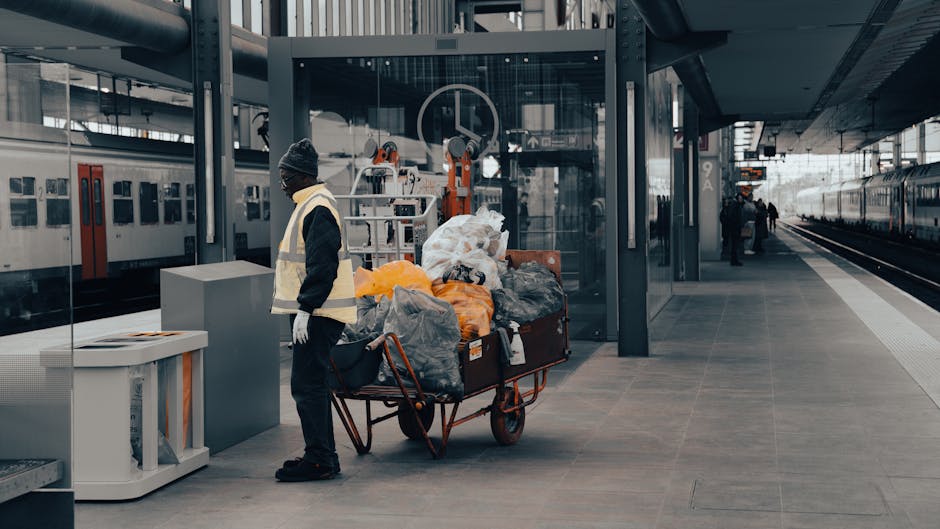 A street cleaner wearing a green safety helmet, face mask, and high-visibility vest with reflective strips is using a mop to clean the pavement near Bromley South station at night. The person is holdi